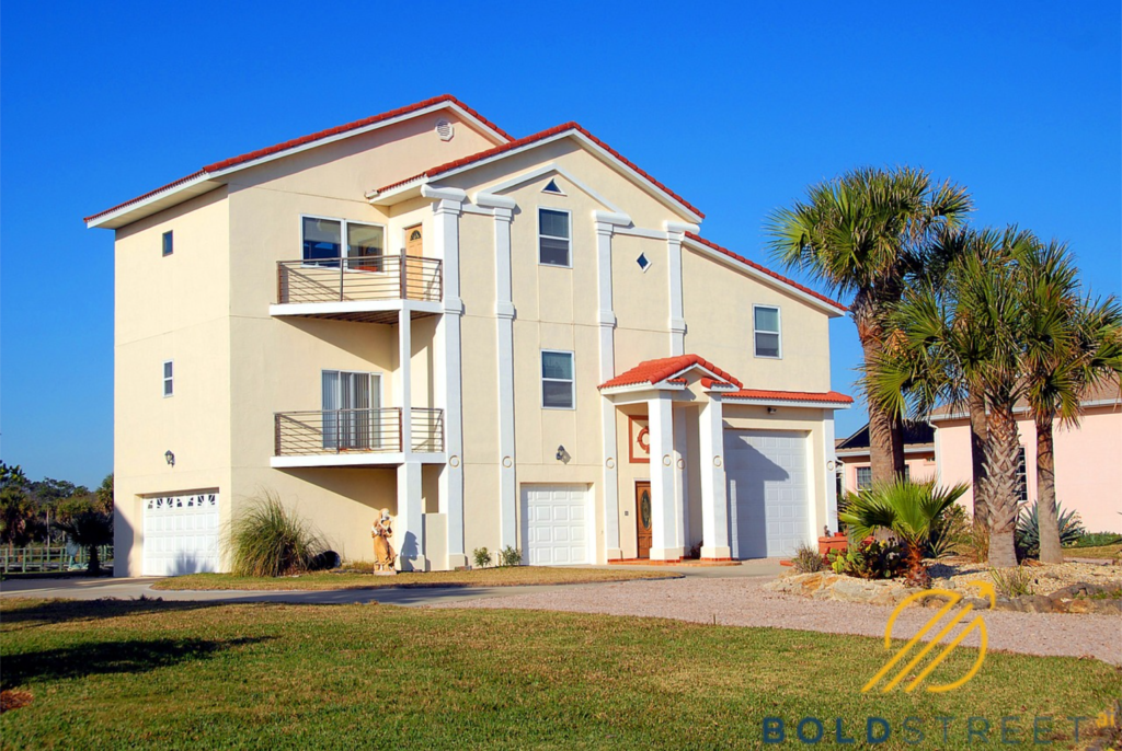 An elegant large white house surrounded by palm trees, featuring windows and a porch, with a driveway leading up to it, set against a clear blue sky.